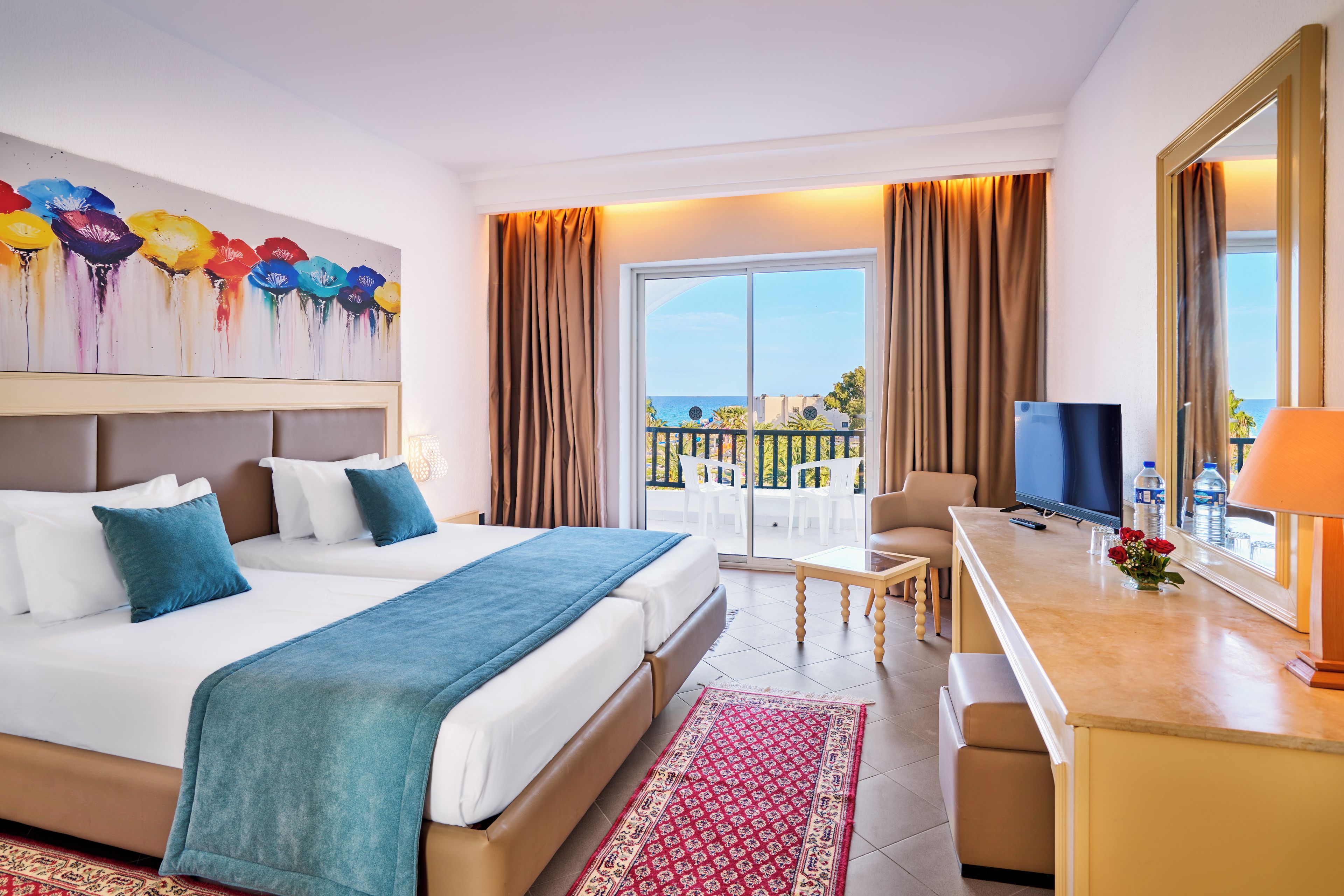 A hotel room with a bed, a desk, a TV, and a balcony with a view of the sea.