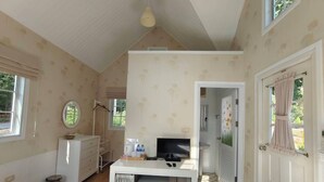 Una habitación con cama, un escritorio con computadora, un televisor y un armario. Hay una ventana y una puerta que da a otra habitación.