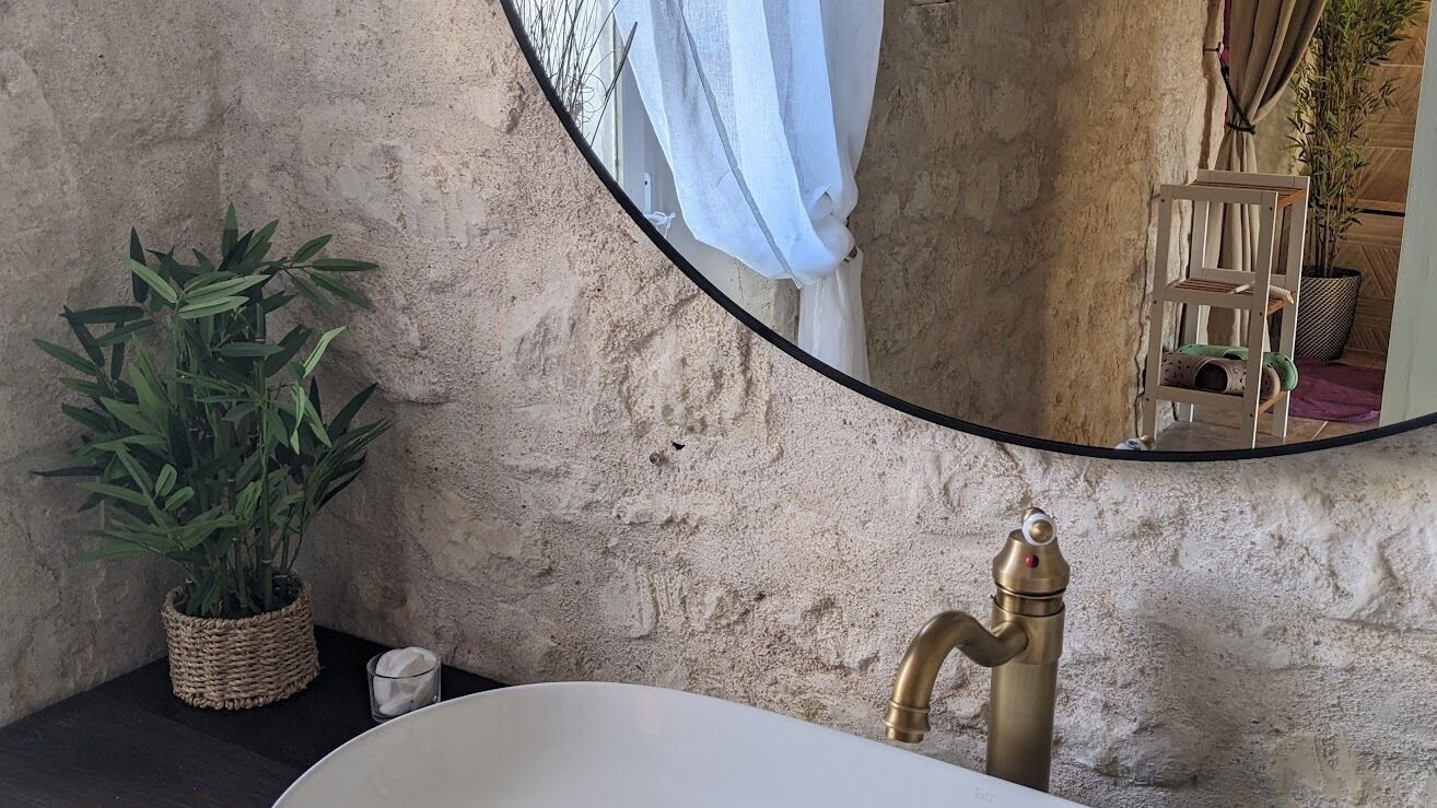 Un baño con un espejo redondo, un lavamanos con grifo dorado, una planta en maceta y una pequeña vela sobre el mostrador.