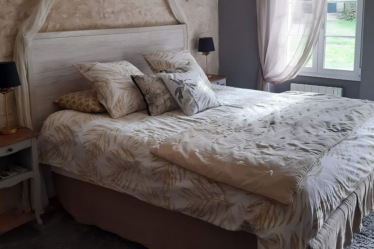 Een slaapkamer met een hemelbed, meerdere kussens, een bedlampje en een raam met gordijnen.