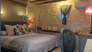 A bedroom with a stone wall, a bed with patterned pillows, a nightstand with a lamp, and a chair with a patterned backrest. - Domaine Les Feuillants  (Crouzilles)