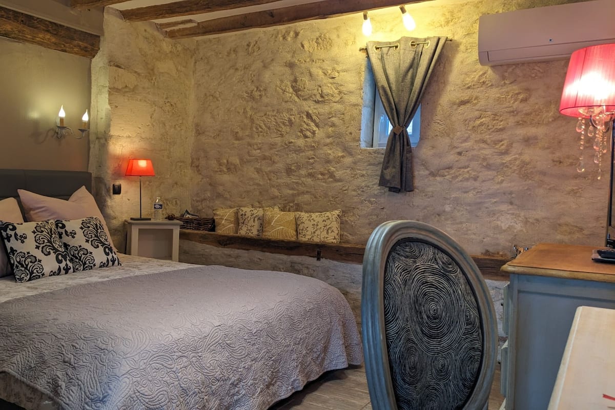 Een slaapkamer met een stenen muur, een bed met gekleurde kussens, een nachtkastje met een lamp en een stoel met een gestreept rugleuning.