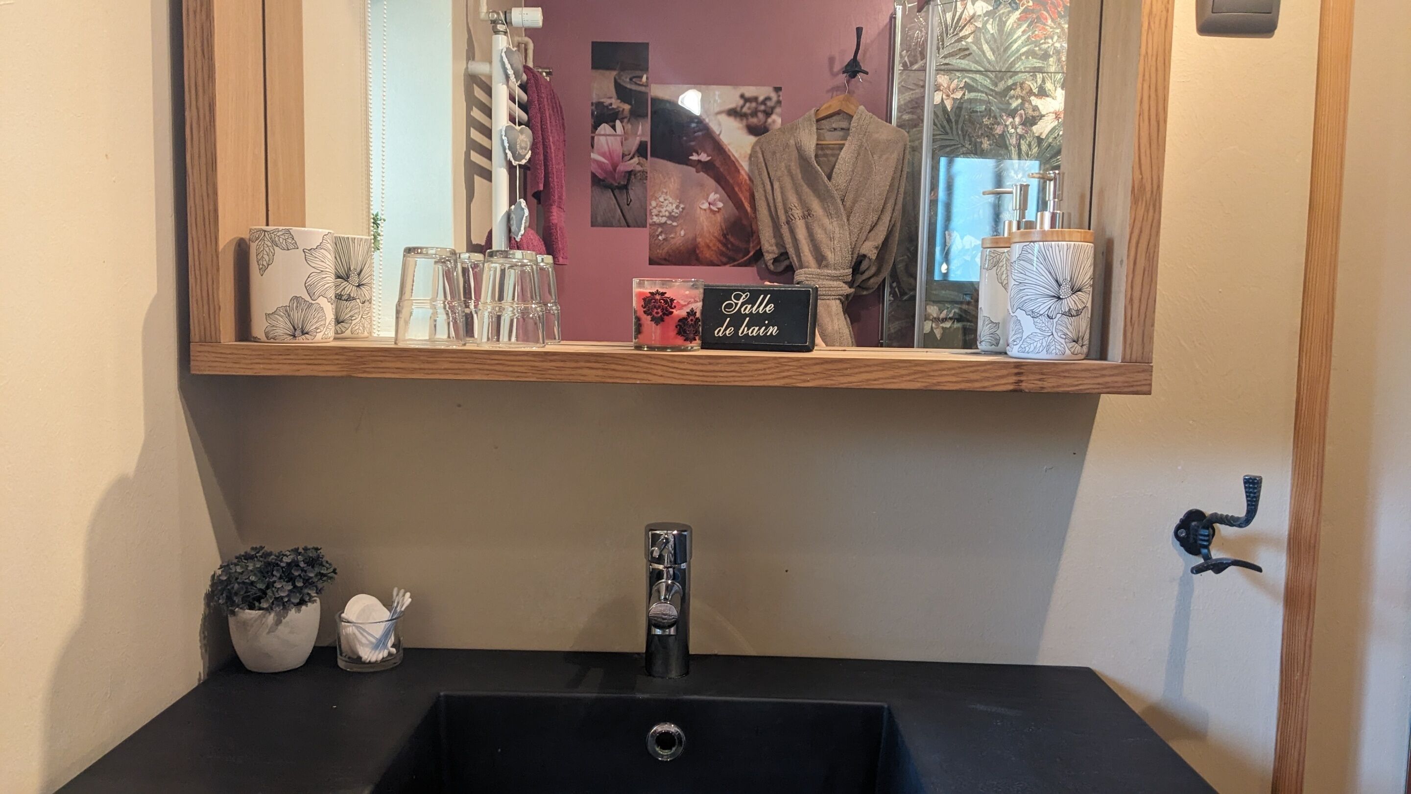 Une salle de bain avec un lavabo noir, un miroir et une étagère au-dessus du lavabo où sont exposés divers objets.