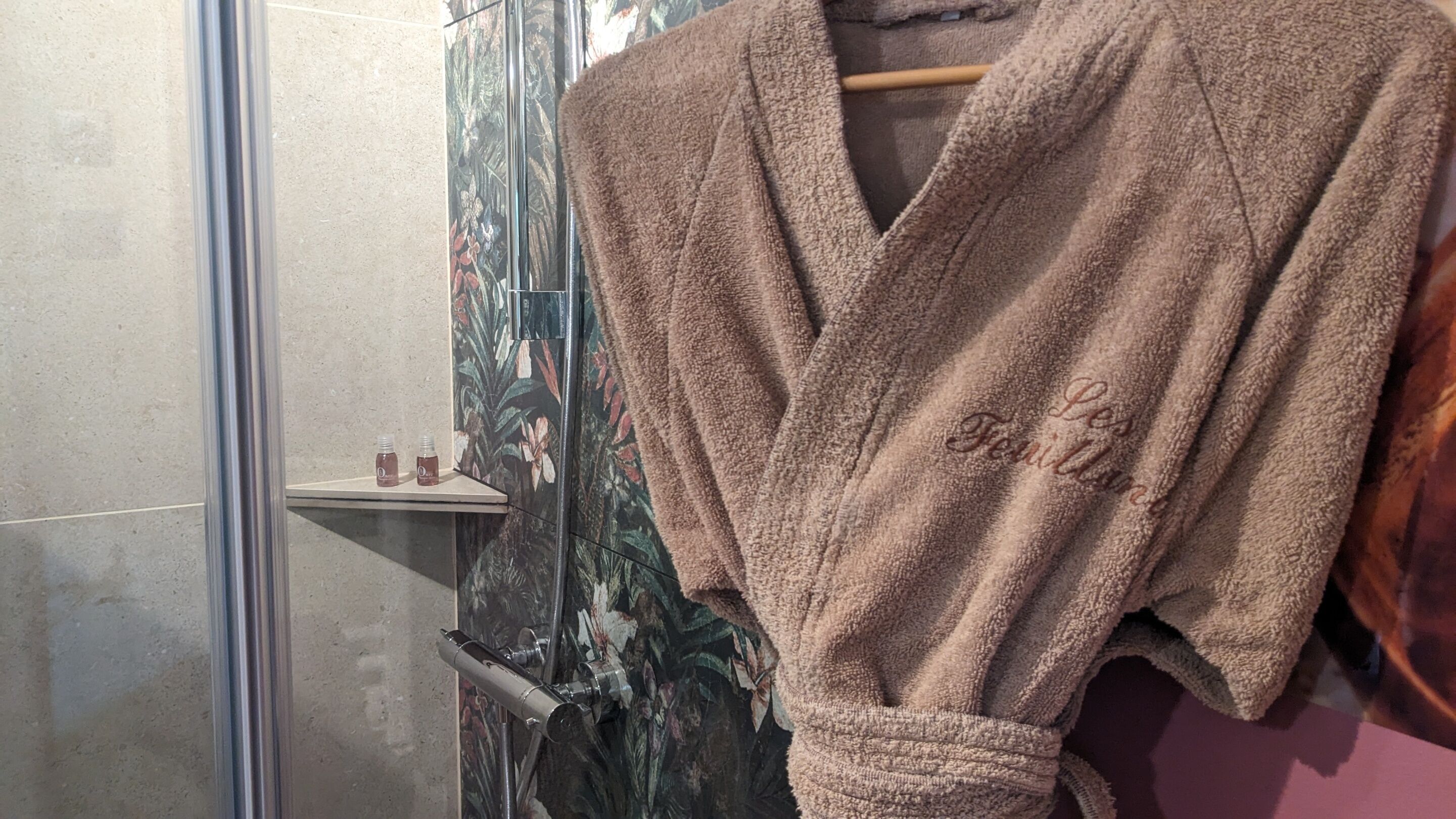 A bathroom with a shower, a towel rack with a robe, and a shelf with two bottles.