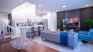 Eine moderne Küche mit Kochinsel, ein Essbereich mit Holztisch und Stühlen, ein Wohnzimmer mit Sofa und blauen Sesseln sowie ein TV-Bereich mit dunkler Holzvertäfelung und einer Pflanze.