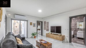 Een moderne woonkamer met een grijze bank, een houten salontafel, een televisie en een badkamer zichtbaar door een open deur.