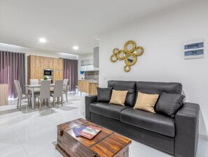 A modern living room with a black leather sofa, a wooden coffee table, and a dining area with a white table and chairs.