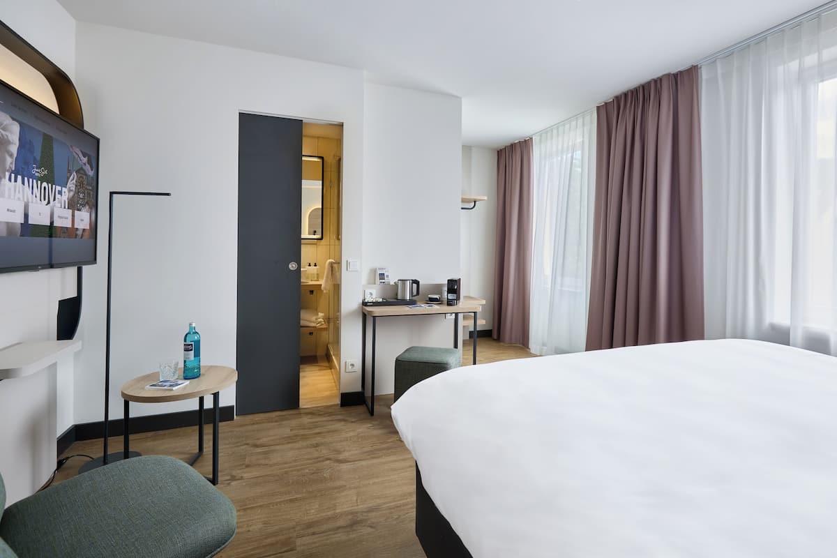 Ein Hotelzimmer mit einem Bett, einem Schreibtisch mit Kaffeemaschine, einem kleinen Tisch mit einer Flasche und einem Fernseher an der Wand.
