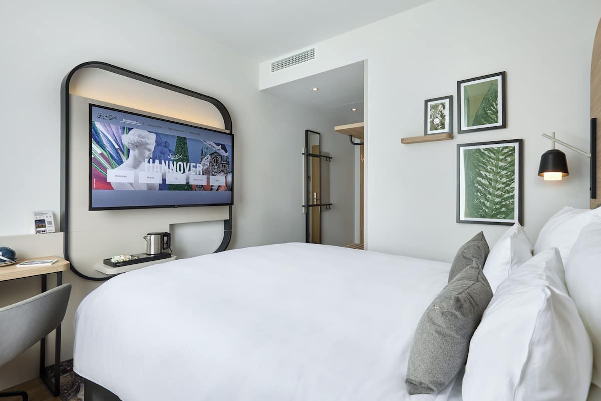 Ein Hotelzimmer mit einem großen Bett, einem Schreibtisch mit Kaffeemaschine, einem Fernseher, der eine Reise-Website anzeigt, und gerahmten Bildern an der Wand.