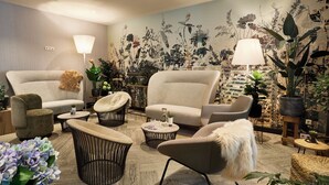 Een moderne woonkamer met een grote bank, fauteuils en een salontafel. Er staan potplanten en er is een muurschildering.