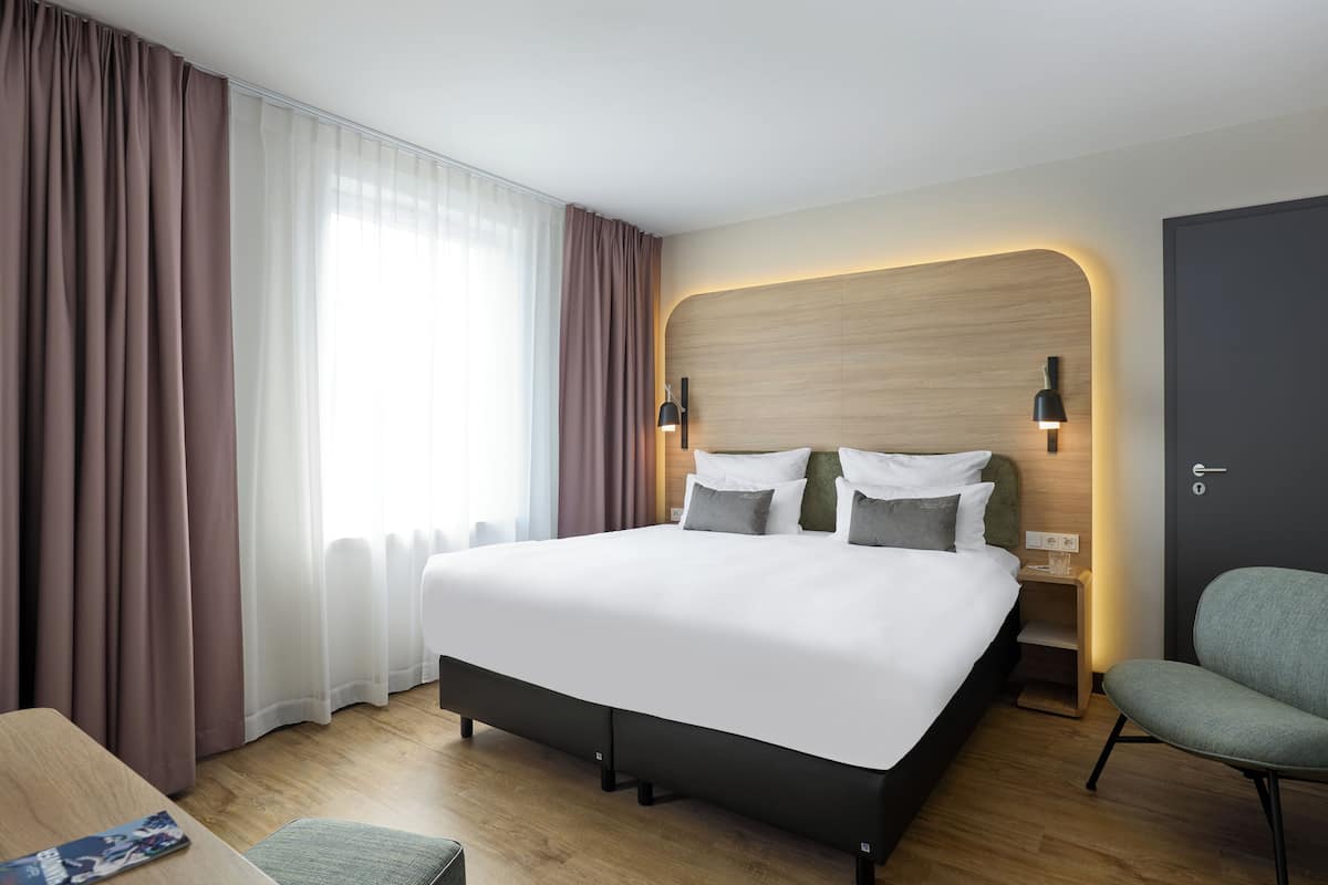 Ein modernes Hotelzimmer mit einem großen Bett, einem Nachttisch, einem Sessel und Blick ins Freie durch transparente Vorhänge.