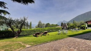 Un área exterior bien cuidada con un sendero pavimentado, sillas de descanso y mesas, rodeada de vegetación y con una montaña al fondo.