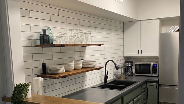 A modern kitchen with white subway tiles, wooden shelves, a stainless steel sink, and a microwave. - Modern 2BD Condo Near TOWN! Fire Pit + Pool Table (Pigeon Forge)