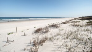 Ein weiter, sandiger Strand mit sanften Wellen, blauem Himmel und vereinzelter Vegetation.