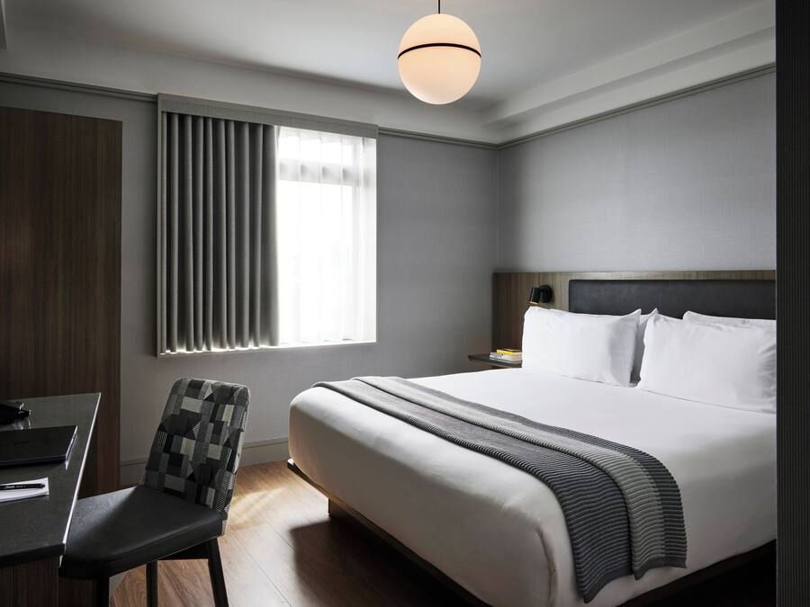 A hotel room with a bed, a desk, a chair, a window with blinds, and a hanging light fixture.