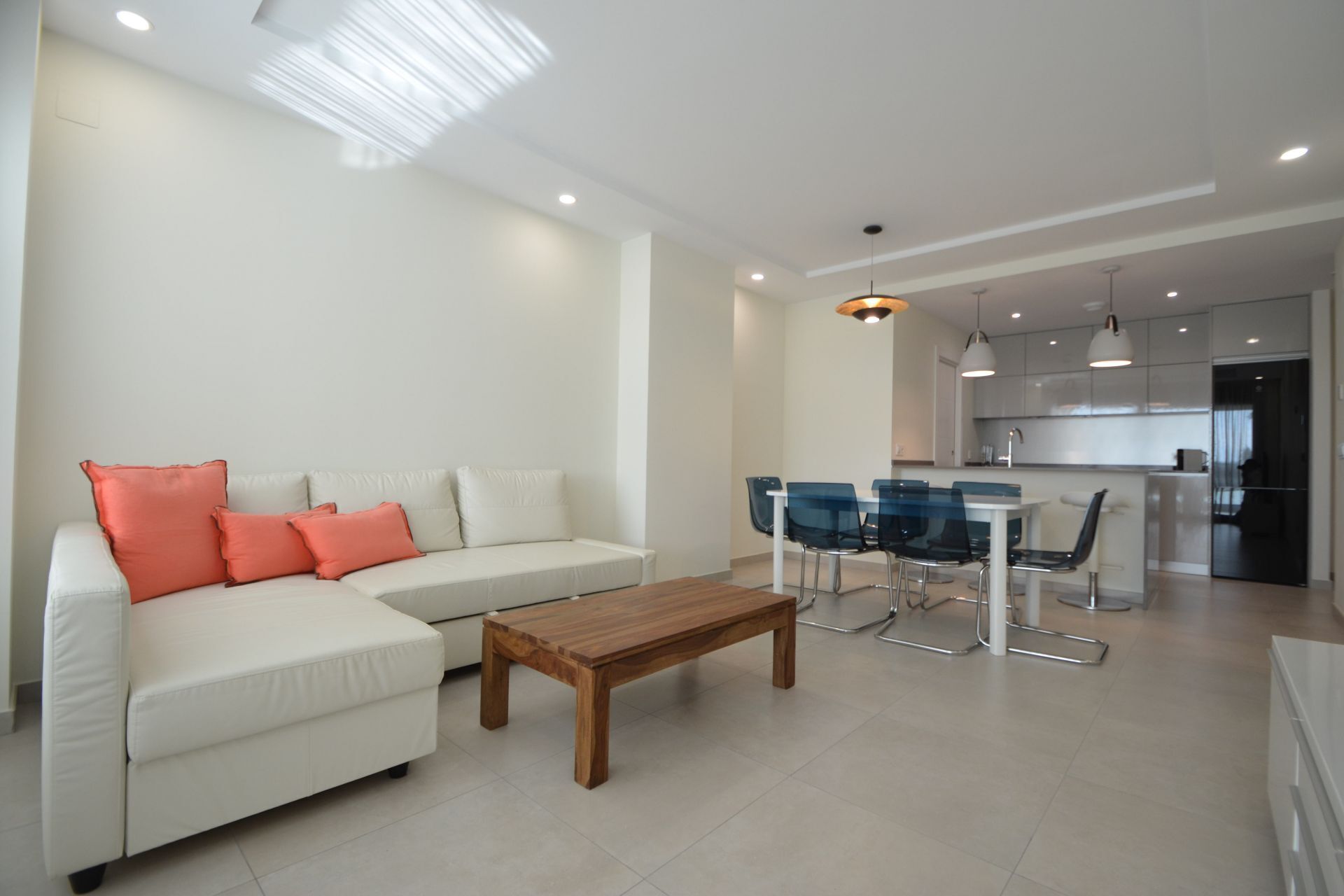 A modern living room with a white sofa, a wooden coffee table, and a dining area with blue chairs and a white table.