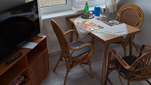 Ein kleines Zimmer mit Fernseher, Tisch mit Stühlen, Fenster mit Blick auf ein Feld und eine Flasche auf der Fensterbank.