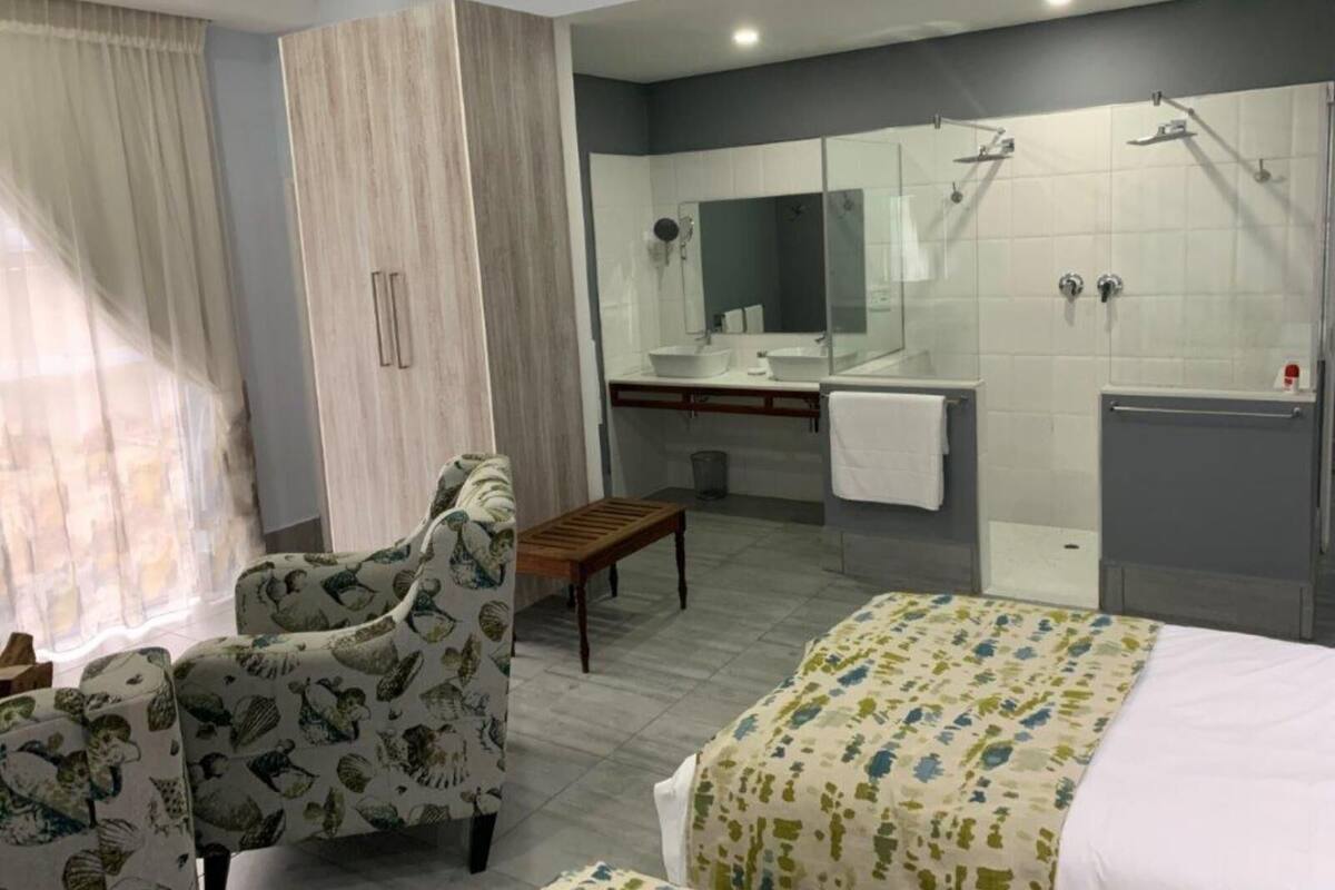 A modern hotel room with a bed, a chair, a small table, a shower area, and a vanity with a mirror.