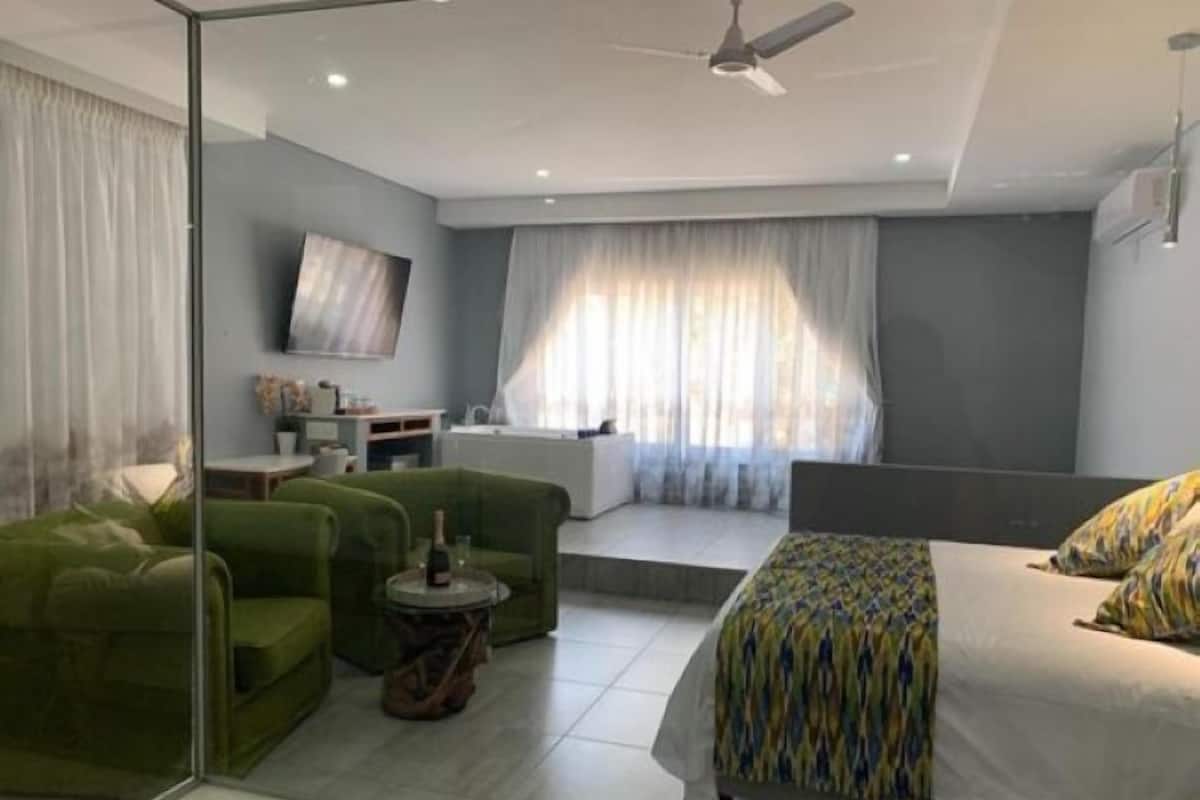 A modern hotel room with a bed, green armchairs, a small table with a bottle, a TV on the wall, and a window with curtains.