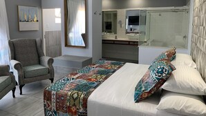 A bedroom with a bed, a chair, a large mirror, a bathroom with a shower, and a painting on the wall. - Ecolux Boutique Hotel Mozambique (Marracuene)