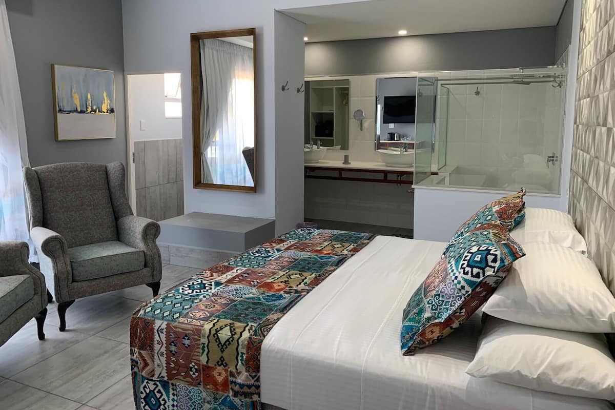 A bedroom with a bed, a chair, a large mirror, a bathroom with a shower, and a painting on the wall.