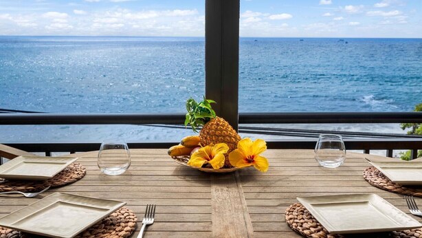 A table set for two with a tropical fruit bowl, a glass of water, and a view of the ocean.