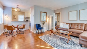A spacious living room with a leather sofa, a glass coffee table, and a patterned rug. There is a dining area with a glass table and chairs, and a kitchen in the background.