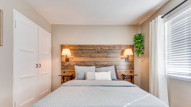 A bedroom with a wooden headboard, two bedside lamps, a bed with grey bedding, and a window with white blinds.
