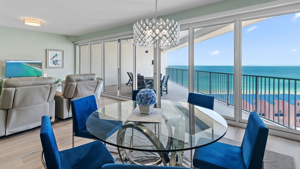 Een ruim balkon met een glazen tafel en blauwe stoelen, met uitzicht op een strand.