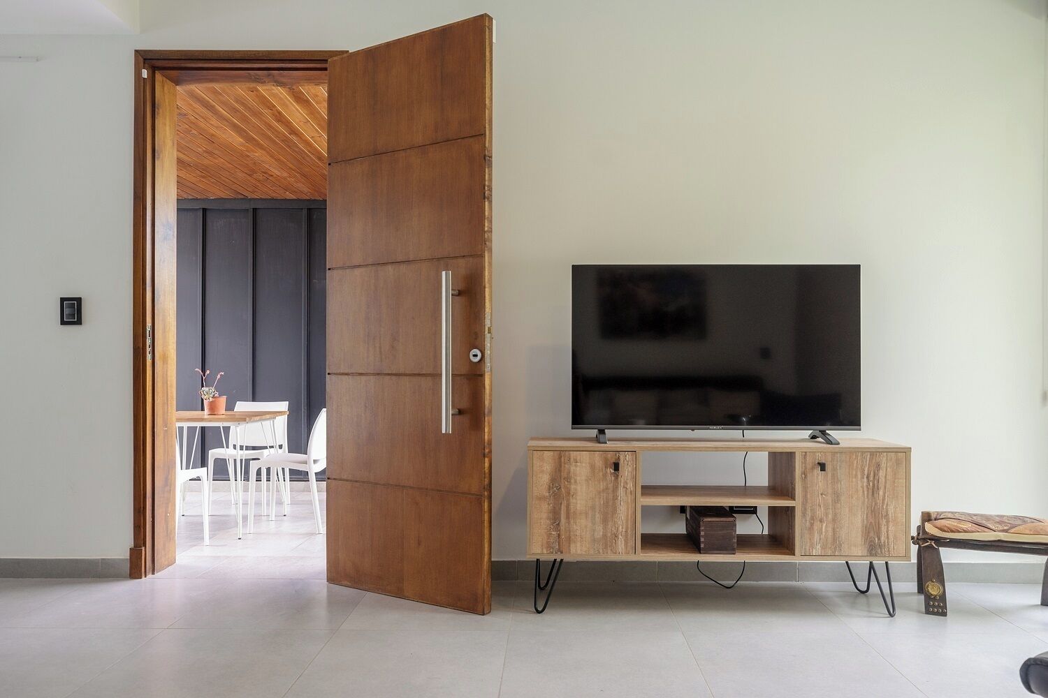 A modern living room with a wooden door, a flat-screen TV on a wooden stand, and a dining area in the background.