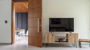 A modern living room with a wooden door, a flat-screen TV on a wooden stand, and a dining area in the background.