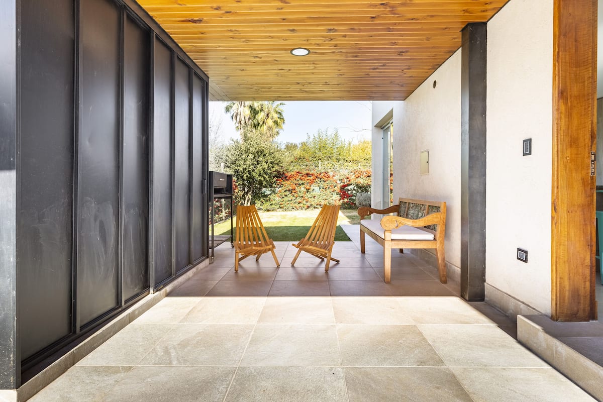 Eine offene Terrasse mit zwei Holzstühlen, einer Bank und Blick auf einen Garten mit roten Blumen und einer Palme.