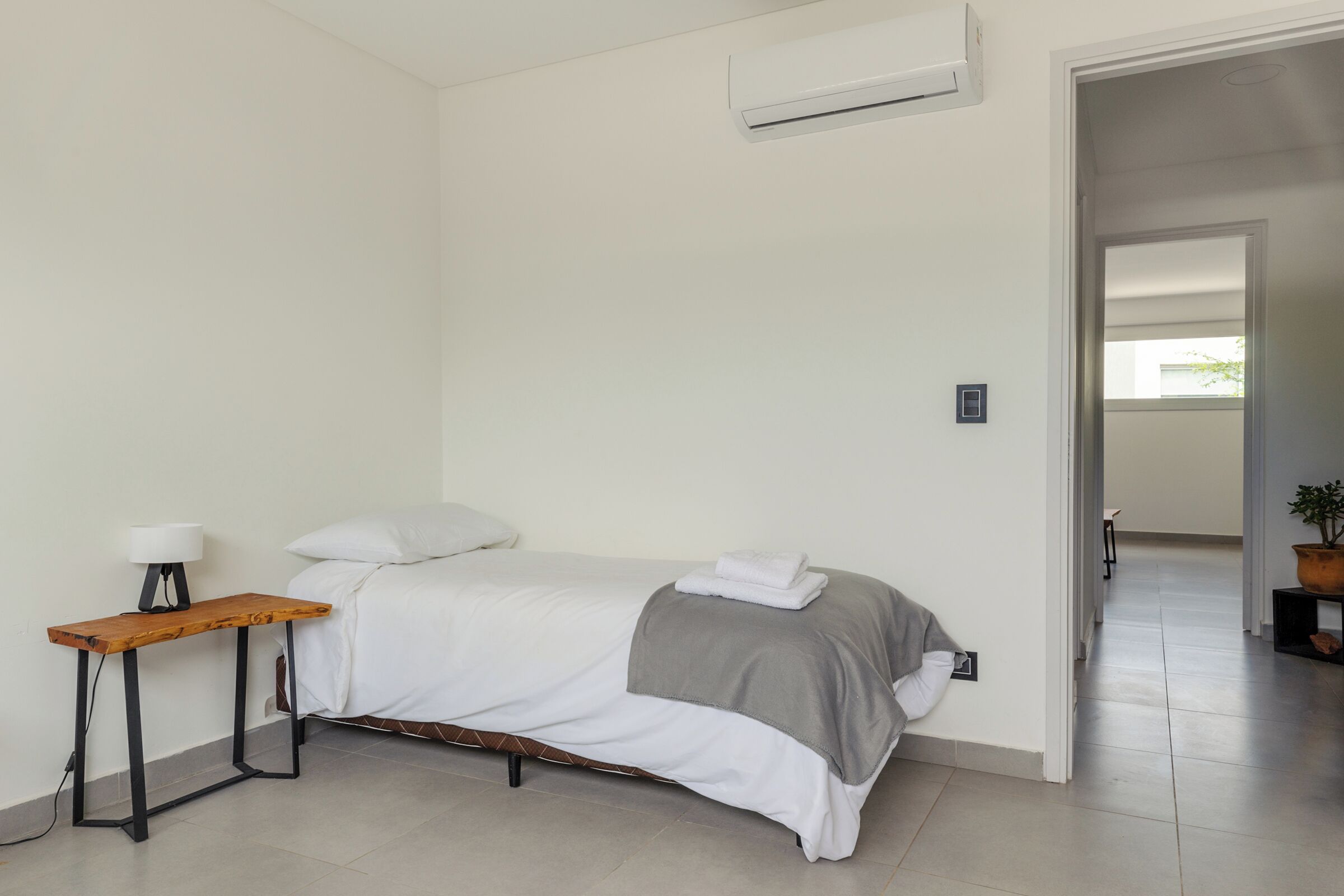 Un dormitorio ordenado con cama, mesita de noche, lámpara y una planta en maceta.