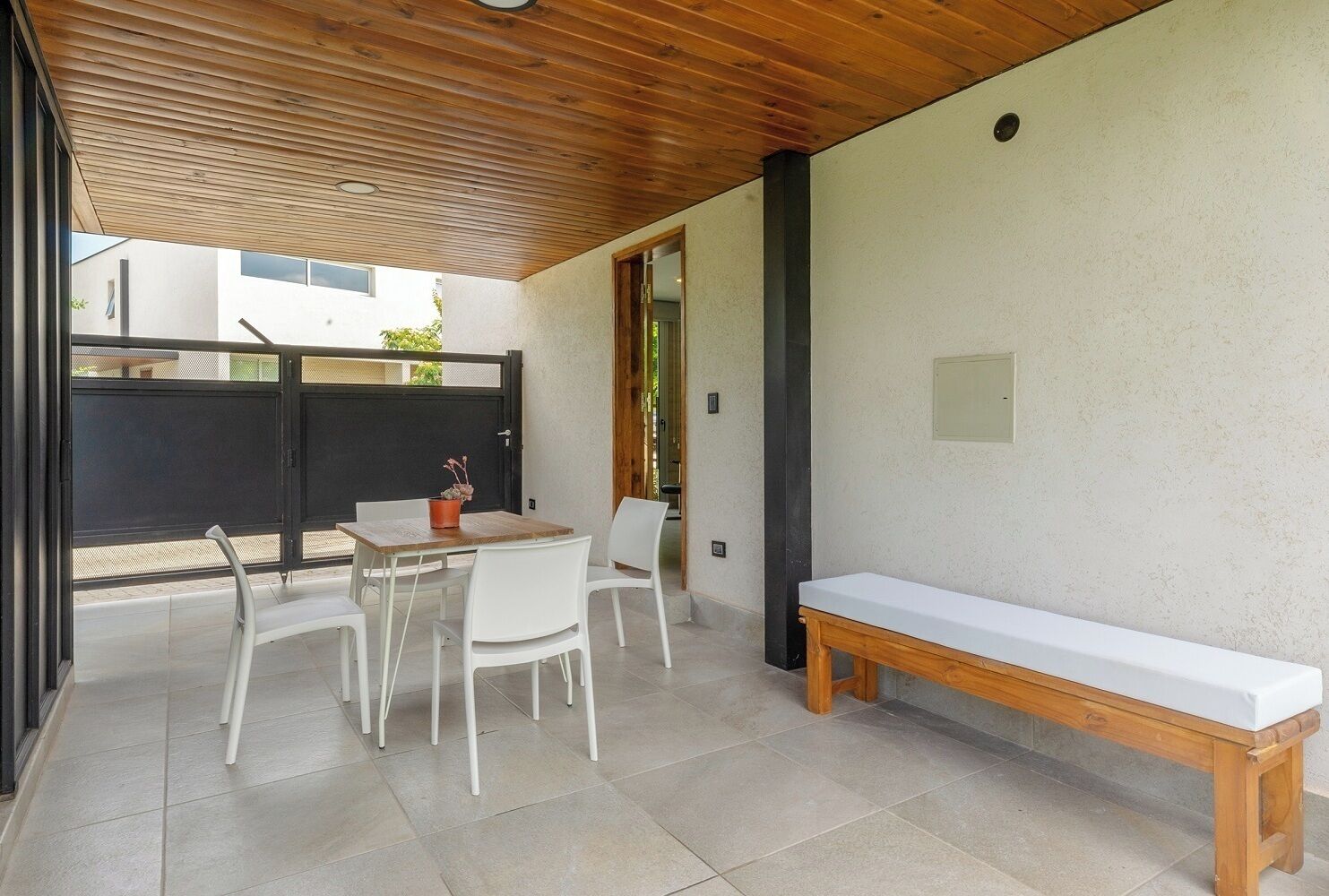 A modern outdoor patio with a wooden table and chairs, a white bench, and a view of a building through a black gate.