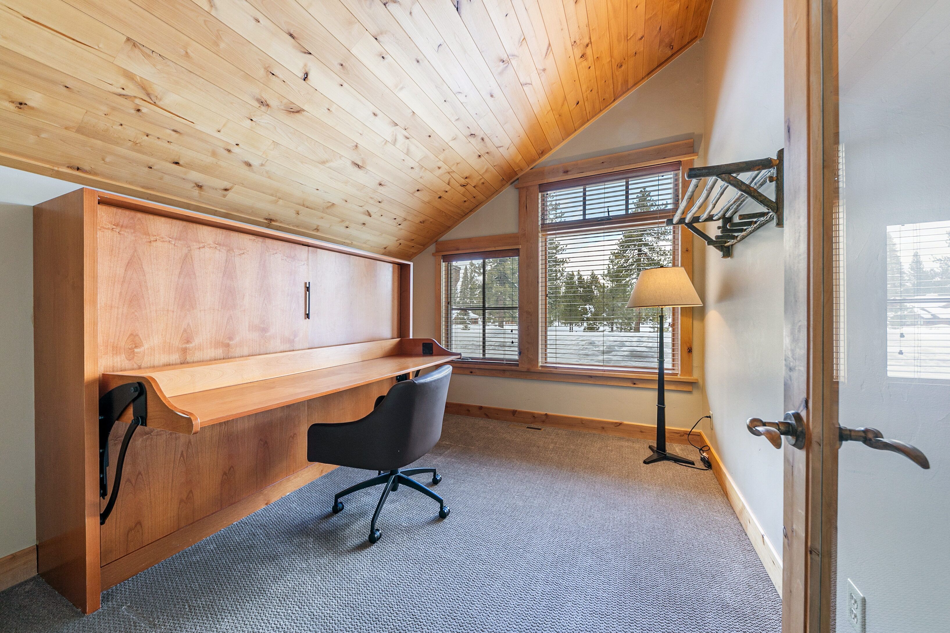 A room with a sloped ceiling, a wooden desk, a black office chair, a standing lamp, and a window with blinds.