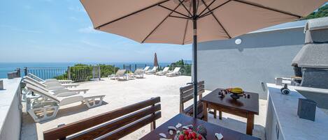 En terrasse ved stranden med et bord, stole og en parasol med udsigt over havet.