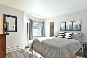 Ein Schlafzimmer mit einem Bett, einem großen Fenster mit Vorhängen, einem Spiegel und drei gerahmten Bildern an der Wand.