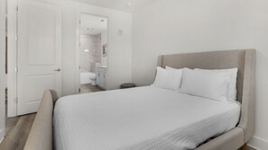 Un dormitorio con una cama, una mesita de noche, una puerta y un baño visible al fondo.