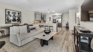 Uma sala de estar moderna com sofá, mesa de centro e banquetas de bar. Cozinha com geladeira e fogão visíveis ao fundo.