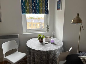 A room with a round table, a chair, a radiator, a window with a view of buildings, a floor lamp, and a decorative item on the table.