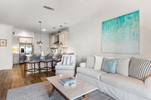 A modern living room with a sofa, a coffee table with books, a kitchen area with bar stools, and a large abstract painting on the wall.