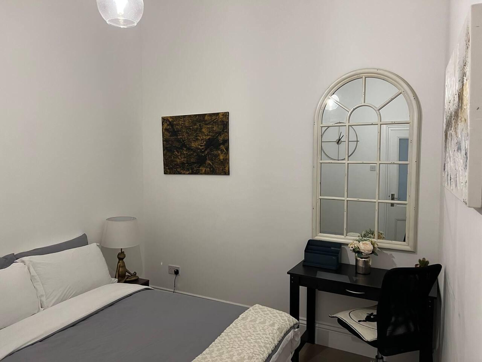 Un dormitorio con cama, escritorio con silla, lámpara, cuadro y ventana con reloj.