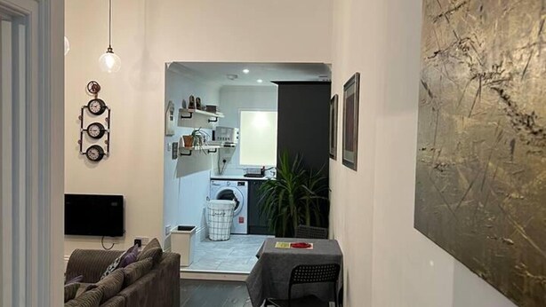 A narrow hallway with a dark wooden floor, a grey sofa, a dining table with chairs, a wall-mounted TV, and a large abstract painting. - Modern and Spacious 2 Bedroom Flat Near Shoreditch (London)