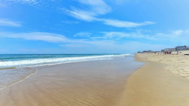 A wide sandy beach with gentle waves, a clear blue sky, and a few buildings in the distance.