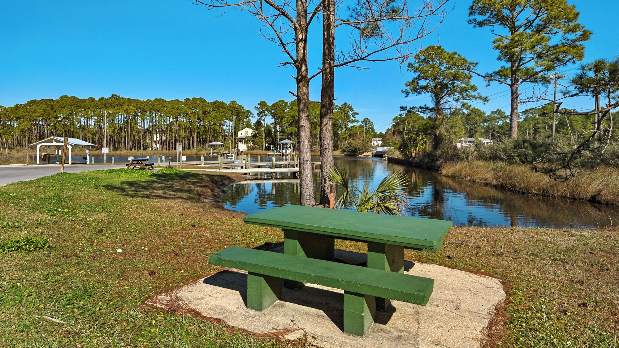 A picnic table by a calm lake with a dock and covered seating area in the background.