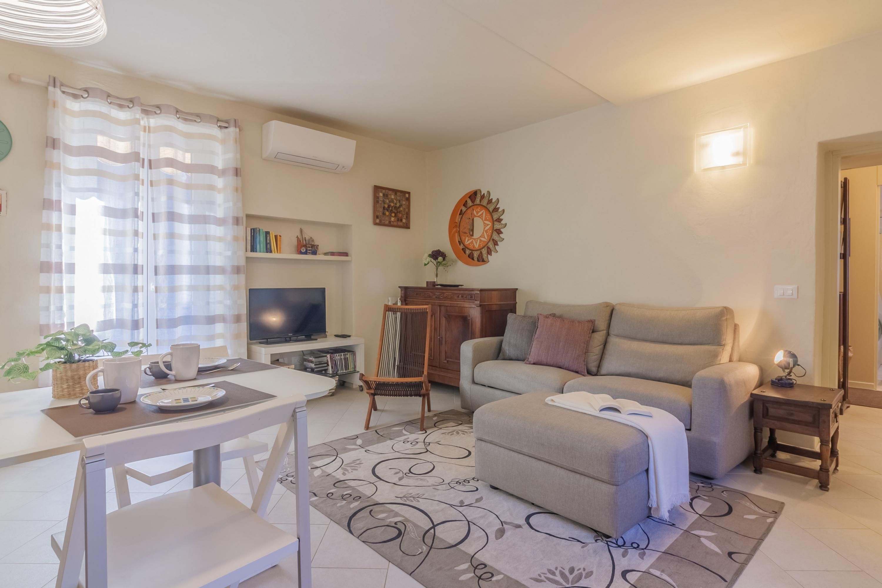 A living room with a sofa, ottoman, and a TV. There is a dining table with chairs and a small table with a lamp.