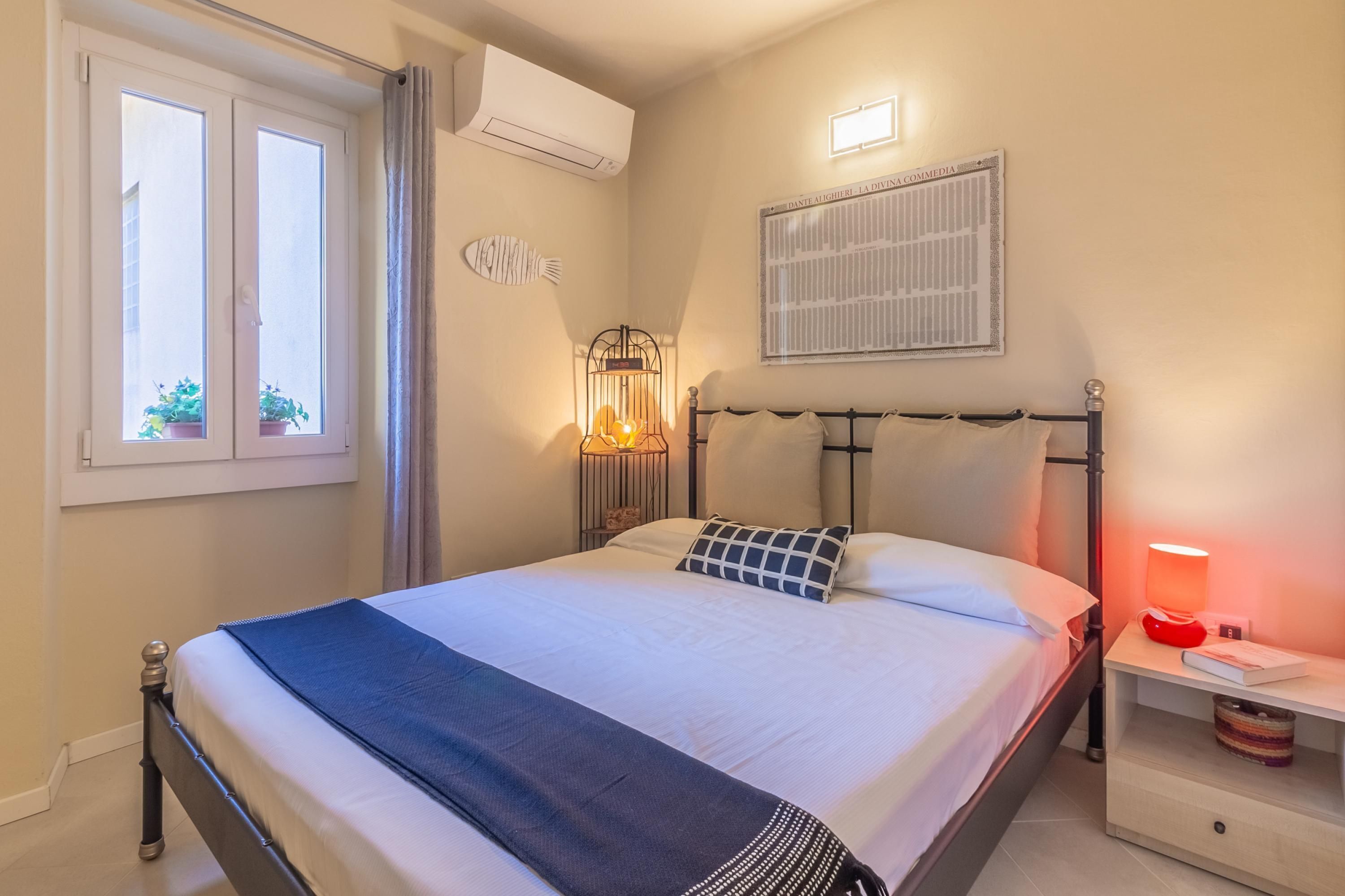 A neatly made bed with a blue throw, a bedside table with a red lamp, and a wall-mounted air conditioner.