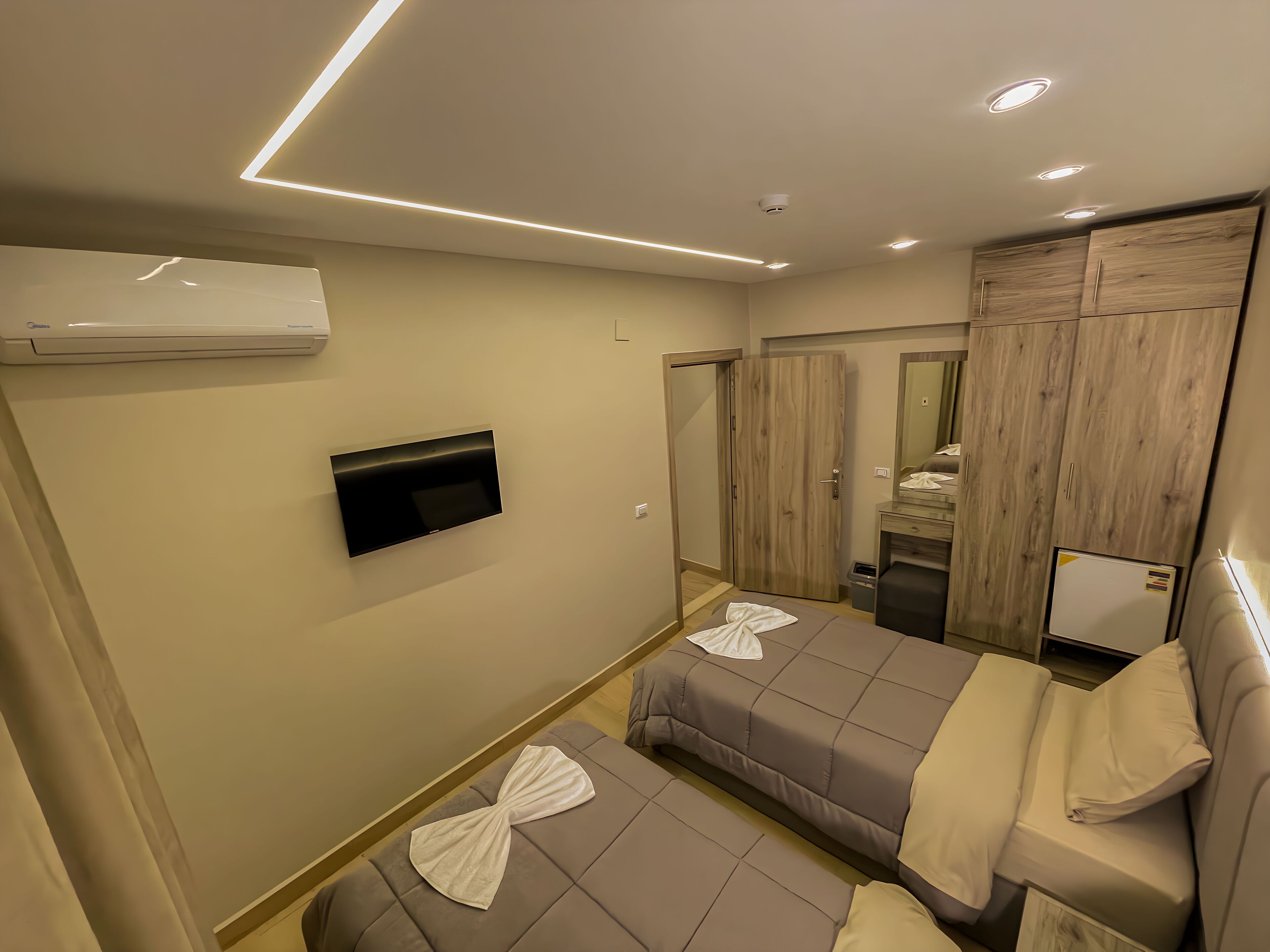 A modern hotel room with a flat-screen TV mounted on the wall, a wooden wardrobe, a mirror, and a bed with towels.