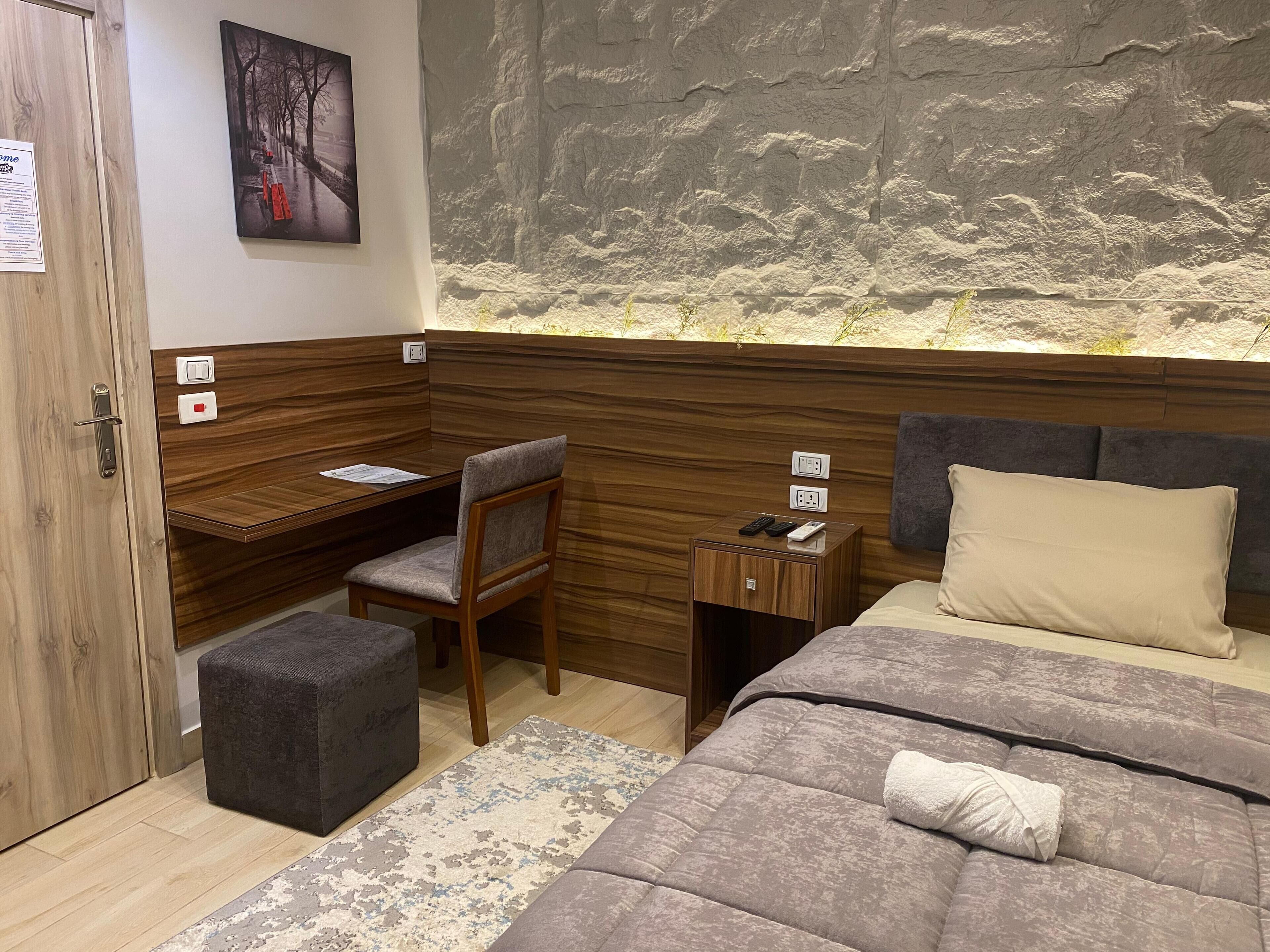 Quarto de hotel compacto com escrivaninha de madeira, uma cadeira, mesa de cabeceira pequena com controle remoto, cama com travesseiro bege e parede texturizada.