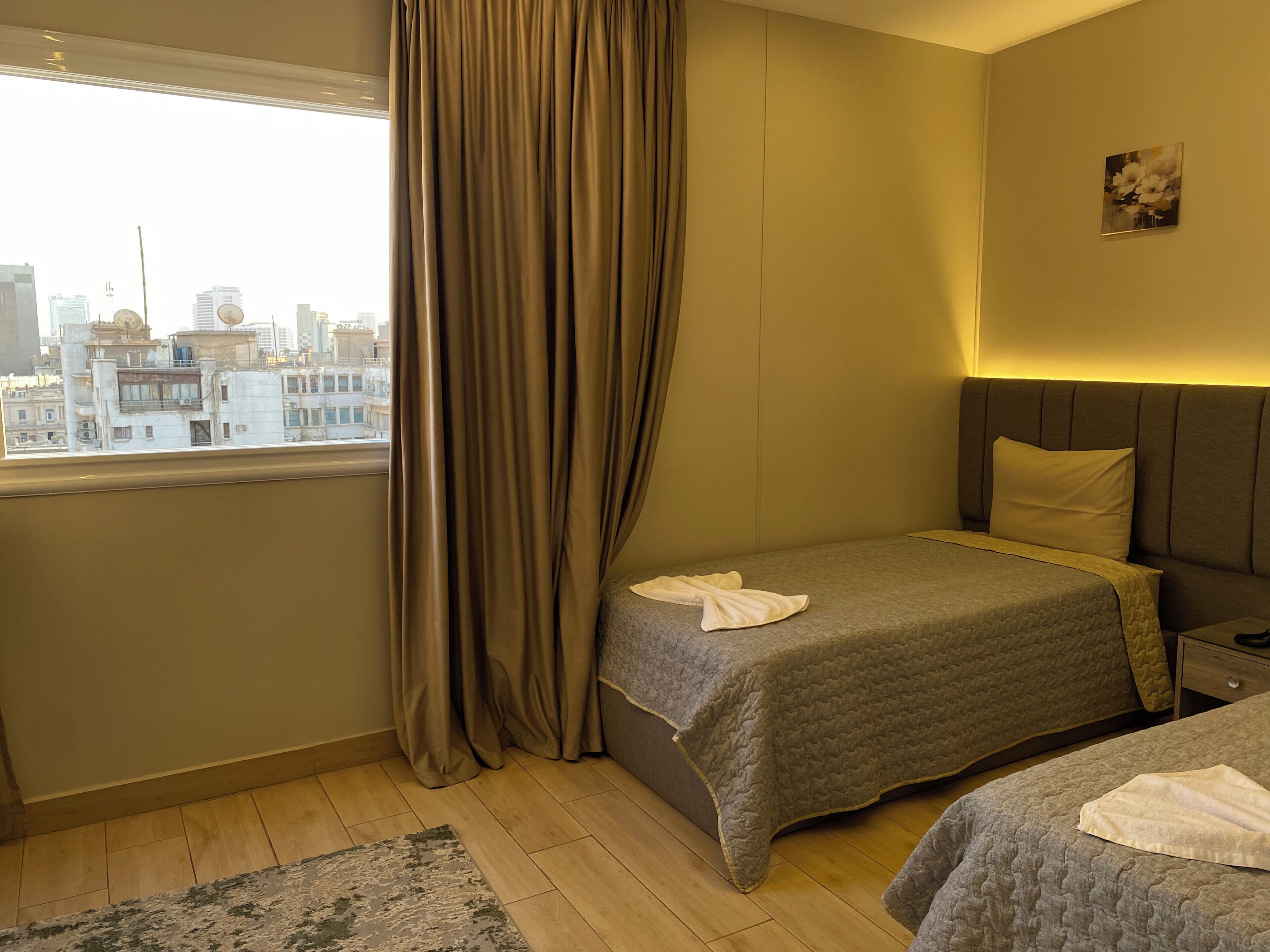 Habitación de hotel con dos camas, una ventana con cortinas, vista a la ciudad y un cuadro pequeño en la pared.